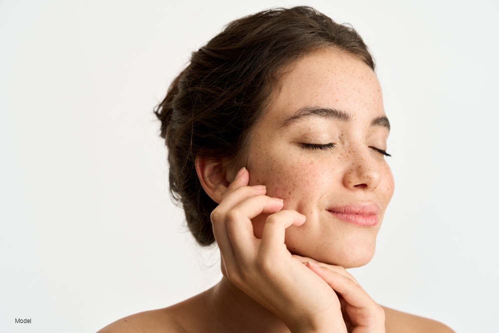 Woman with freckles and smooth skin touching her face and smiling with her eyes closed.
